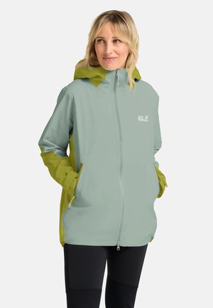 Frau mit blonden Haaren, die eine hellgrüne und olivfarbene Kapuzen-Regenjacke trägt und die Hände in den Taschen hat, stehend vor einem schlichten weißen Hintergrund.