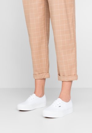 Nogi w beżowych, w kratę spodniach podwiniętych przy kostkach oraz białych sneakersach Vans na platformie na jasnoszarej podłodze.