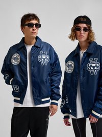 Deux jeunes adultes portant des vestes varsity bleu marine avec des logos blancs, des lunettes de soleil et des vêtements de rue décontractés posent devant un fond gris uni.