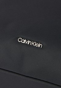 Μαύρος υφασμάτινος ιστός με εξέχον λογότυπο "Calvin Klein" σε ασημί, με λεία επιφάνεια και κομψό σχέδιο.