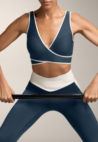 Temno modra športna modrček z belimi detajli in obliko V-izreza, v kombinaciji z usklajenimi visoko pasnimi pajkicami, gladka tekstura, telesu prilegajoča silhueta.