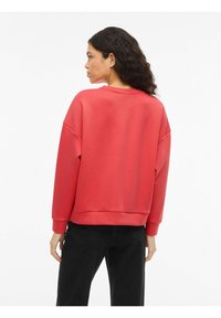 Czerwona bawełniana bluza z opuszczonymi ramionami, ściągaczami na mankietach i u dołu. Gładka faktura, luźny krój. Widok z tyłu ukazuje detale dekoltu i szwów.