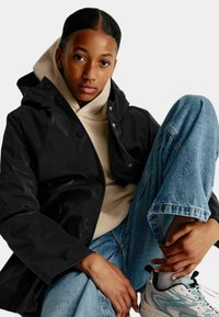 Sort vandtæt jakke med hætte, beige hoodie under, og lyseblå slidte jeans. Model iført grå og hvide sneakers.
