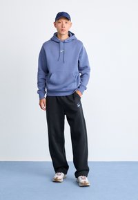Lyseblå hettegenser med frontlomme, kombinert med svarte bukser med logo, og grå joggesko med brune detaljer.
