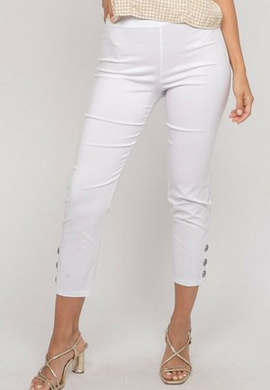 Femme portant un pantalon blanc ajusté et court avec des détails à boutons aux chevilles et des sandales métalliques à talons hauts avec des lanières.