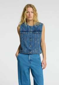 Chaleco de mezclilla en azul claro con cierre de botones, dos bolsillos en el pecho y un diseño corto. Lleva puesto sobre pantalones de mezclilla de tiro alto a juego.