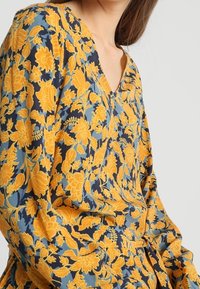 Vestido de manga larga en tejido azul oscuro con un estampado floral en amarillo y azul claro. Presenta un escote en forma de V y detalle fruncido en la cintura.