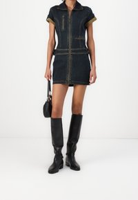 Kvinna i en mörk denimminiklänning med dragkedja framtill, uppvikta ärmar, svarta knähöga stövlar och håller en liten svart handväska.