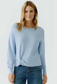 Frau mit schulterlangen blonden Haaren, die ein hellblaues Langarmoberteil trägt, das in der Taille gebunden ist, und blaue Jeans trägt, stehend vor einem weißen Hintergrund.