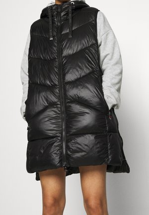 Osoba nosząca bezrękawnik czarny, ocieplany z kapturem na jasnoszarym swetrze, ręce w kieszeniach kamizelki, stojąca na jednolitym tle.