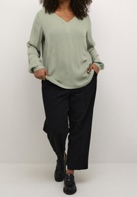 Helles grünes Langarmoberteil mit V-Ausschnitt und subtiler Textur, kombiniert mit locker sitzenden schwarzen Hosen und schwarzen Slippern.
