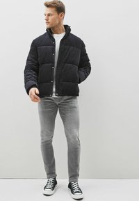 Jachetă puffer de culoare bleumarin închis, cu un design matlasat, butoane cu clips și guler înalt. Blugi din denim gri și adidași negri completează aspectul.