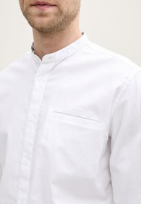 Camicia bianca in cotone con colletto alla mandarino, patta frontale e un'unica tasca sul petto. Il tessuto ha una fine trama e una finitura liscia.