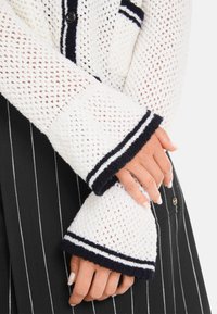 White knitted cardigan with a loose weave, featuring navy and white striped cuffs and a front button detail. Black striped skirt visible below.