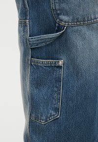 Κοντινό πλάνο μπλε τζιν με πλαϊνή τσέπη εργαλείων, βρόχο από πάνω και ενισχυμένη ραφή με λεπτομέρεια με νίκελ.