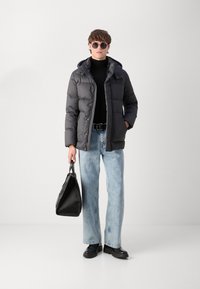 Puhutud hall välimüts koos kapuutsiga, mustides turtleneck, helesinised sirged teksad, must kott ja mustad kingad. Ümmargused prillid.
