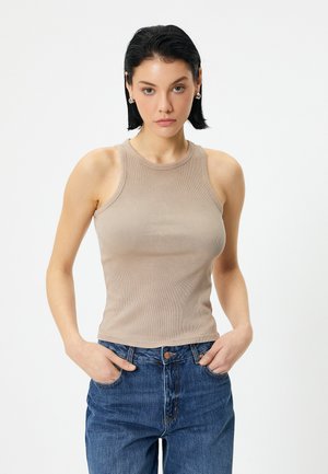 Nude ribbed tank top dengan leher bulat dan desain tanpa lengan, dipadukan dengan celana jeans denim biru berpinggang tinggi, tangan di saku.