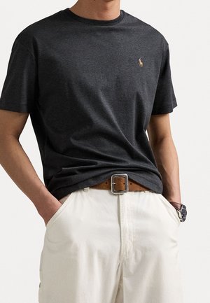 Personne portant un t-shirt gris foncé à manches courtes avec un petit logo brodé, rentré dans un pantalon blanc avec une ceinture marron et une montre au poignet.
