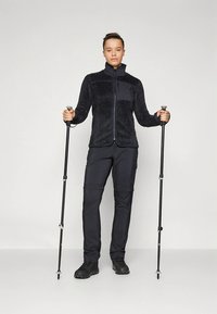 Flis jakna temno sive barve z zadrgo spredaj, teksturirano tkanino in enobarvnim sivim panelom. Črne hlače in pohodniške palice dopolnjujejo zunanjo opravo.