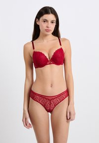 Roter Spitzen-BH mit gewellten Kanten und floralen Stickereien, kombiniert mit einem passenden transparenten Slip, der ähnliche Muster aufweist und einen tief sitzenden Schnitt hat.