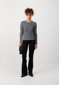 Schwarz-weiß gestreiftes Langarmshirt kombiniert mit weit geschnittenen schwarzen Hosen. Die Männermodel hält eine kleine schwarze Handtasche. Einfaches, figurbetontes Design.