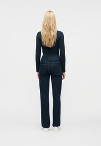 Γυναίκα με μακριά ξανθά μαλλιά φοράει μπλε μακρυμάνικη μπλούζα, σκούρα μπλε τζιν με ίσι α γραμμή και λευκά αθλητικά παπούτσια, στέκεται με την πλάτη προς τα εμπρός σε λευκό φόντο.