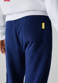 Dunkelblaue Sweatpants mit strukturiertem Finish, ausgestattet mit einer Gesäßtasche und einem kleinen gelben Markenetikett auf der Rückseite.