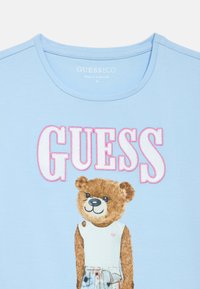 Ανοιχτό μπλε βαμβακερό T-shirt με γραφικό αρκούδας που φοράει λευκό πουκάμισο και σορτς με σχέδιο, με την επιγραφή "GUESS" σε ροζ χρώμα από πάνω.