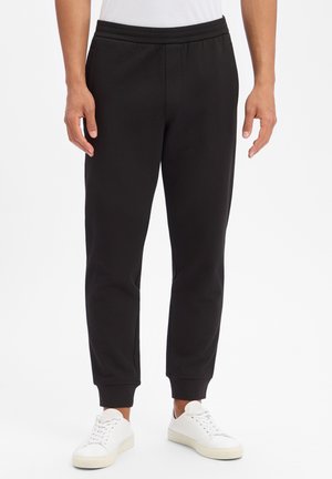 Man die zwarte joggerbroeken en witte sneakers draagt, staand tegen een effen witte achtergrond.