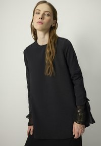Black long-sleeve top with a round neckline, featuring sheer, flared cuffs and a soft texture. Includes a small chest pocket detail.
