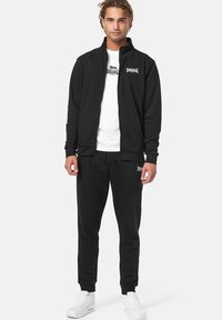 Chaqueta negra con cremallera y dos bolsillos frontales, combinada con pantalones jogger negros a juego. Presenta un detalle de logo blanco en el pecho y en el lateral. Zapatillas blancas.