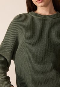 Pull en tricot vert olive avec une texture côtelée, un col rond et des épaules tombantes, affichant une coupe décontractée.