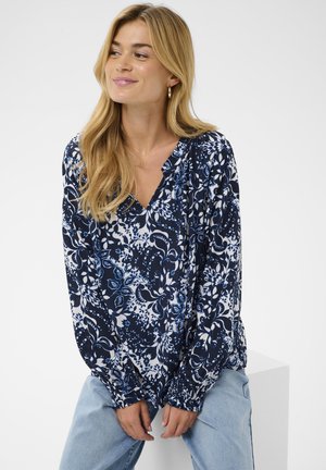 Tumši zila ziedu blūze ar V veida izgriezumu, garām piedurknēm un savāktām aprocēm. Audums ir ar baltu un zilu botānisku rakstu.