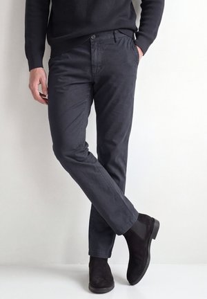 Person trägt dunkelgraue Hose und schwarze Chelsea-Stiefel aus Wildleder, steht mit einem Bein vor das andere gekreuzt vor einem weißen Hintergrund.