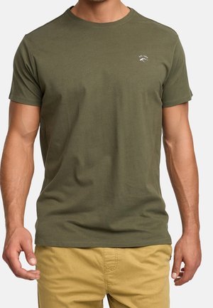 Homme portant un t-shirt vert olive uni à manches courtes avec un petit logo blanc sur la poitrine et un pantalon jaune moutarde, debout devant un fond blanc.