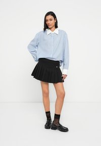 Ανοιχτό μπλε ριγέ πουκάμισο με λευκό γιακά, συνδυασμένο με μαύρη φούστα με πιέτες. Η εμφάνιση ολοκληρώνεται με μαύρα γυαλιστερά παπούτσια και διάφανες μαύρες κάλτσες.