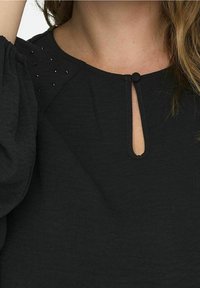 Blusa negra con un escote de ojal en el cuello, tejido texturizado y hombros adornados con pequeños detalles negros.