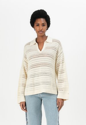 Knit pullover in cream, featuring a collared V-neck, long sleeves, and a textured pattern with openwork details. Paired with light blue jeans.