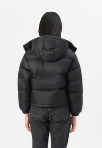 Personne portant une doudoune noire avec capuche, vue de dos, associée à un jean denim gris foncé sur un fond uni.