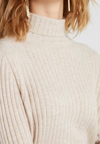 Suéter de cuello alto acanalado beige con un collar alto, fabricado en un tejido suave, que presenta un patrón texturizado y una silueta ajustada.