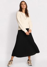 Strikket cremefarvet sweater med kabelmønster, kombineret med en sort plisseret midiskirt og beige sportssko, hvilket skaber et lagdelt outfit.