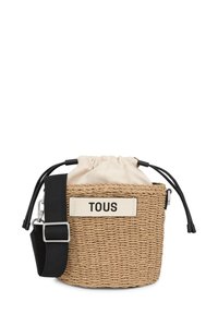 Borsa a secchiello in paglia intrecciata con un bordo in tessuto crema, tracolla nera regolabile e un'etichetta bianca con la scritta "TOUS". Superficie testurizzata.
