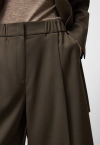 Ciemnobrązowe spodnie w kant z dopasowaną talią, o gładkiej teksturze i subtelnym detalem plisowania, połączone z jasnobrązową bluzką.