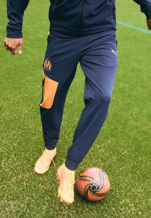 Dunkelblaue Jogginghosen mit orangefarbigen Einsätzen, mit einem Logo und einer Reißverschlusstasche. Helle gelb-orangefarbene Fußballschuhe. Ein gemusterter Fußball auf Gras.