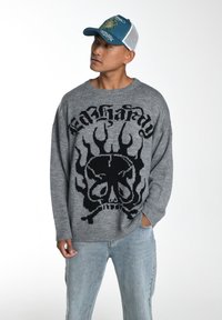 Grau gestrickter Pullover mit schwarzem Schädel- und Flammenmuster. Getragen mit hellblauen, ausgebleichten Jeans und einer blau-weißen Kappe. Lässige Passform.