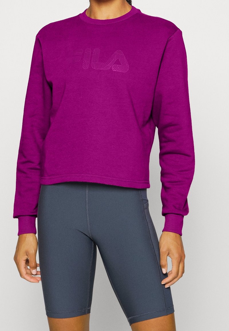 Fioletowy krótki sweatshirt z teksturowanym logo z przodu, w połączeniu z szarymi, prążkowanymi szortami kolarskimi o dopasowanym kroju.