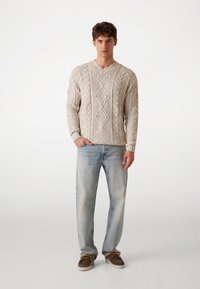 Helle beige Kabelstrickpullover mit V-Ausschnitt, der ein strukturiertes Muster aufweist. Kombiniert mit hellblauen Jeans im geraden Schnitt und braunen Schuhen.