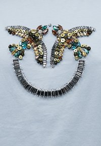 Δύο συμμετρικά σχήματα πεταλούδας φτιαγμένα από πολύχρωμες χάντρες και παγιέτες πάνω από μια καμπύλη σειρά ορθογώνιων σκούρων κρυστάλλων σε ανοιχτό ύφασμα.