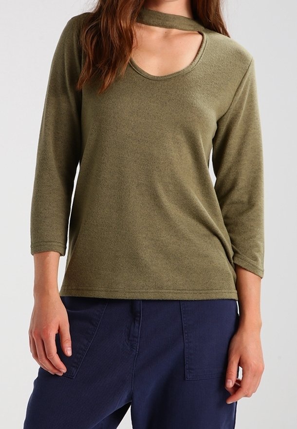 Femme portant un haut à manches longues vert olive avec un col choker et un pantalon bleu marine sur un fond clair uni.