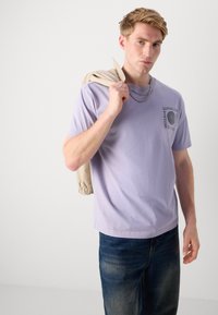 Lyselilla kortermet t-skjorte med grafisk trykk på venstre bryst, parret med blå denimjeans. Lys beige jakke lagt over skulderen.
